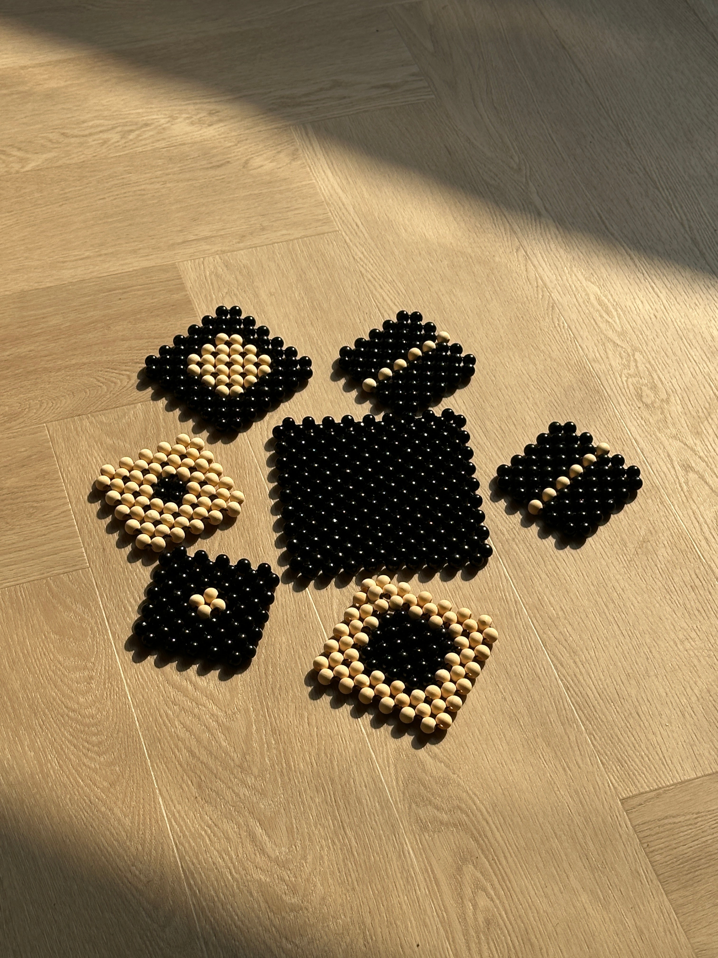 Seven square floor mats made of small wooden beads, arranged on a light wood herringbone floor. The mats alternate between solid black and black-and-cream patterns.
