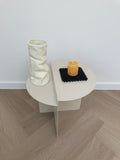 A modern wall-mounted toilet paper holder with a white oval backplate, a white paper tube holder, a black textured mat, and a small yellow holder, shown against a white wall and light wood herringbone floor.